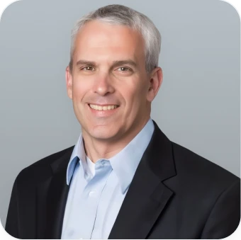 Smiling man in a blazer and light blue shirt against a plain background.