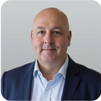 Bald man in a navy suit and light blue shirt, smiling against a plain gray background.
