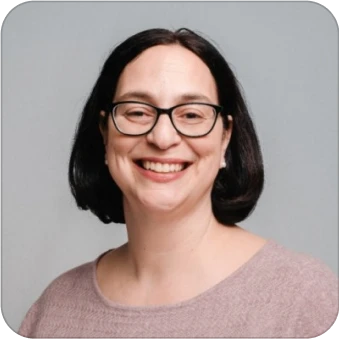 Smiling person with glasses and short dark hair, wearing a light purple sweater against a gray background.